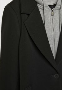 Manteau noir cintré superposé sur un sweat à capuche gris clair zippé avec une fermeture éclair métallique visible et une capuche.