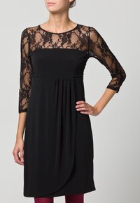 Musta mekko, jossa on kukkakuvioinen pitsi kolmen neljänneksen hihoissa, rypytetty torso ja löysä malli. Hameessa on drapadoitu muotoilu, ja se ulottuu polven ylle.