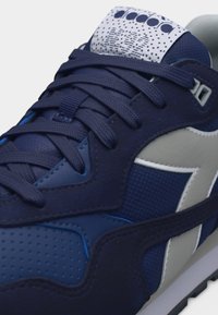 Σκούρο μπλε αθλητικό παπούτσι με λευκό λογότυπο στο πλάι, υφασμάτινες και δερμάτινες επιφάνειες, και σκούρα μπλε κορδόνια δεμένα πάνω από μια διάτρητη μύτη.