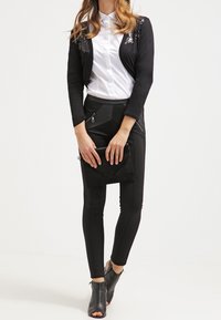 Donna con un blazer nero cropped con dettagli in paillettes, camicia bianca con bottoni, pantaloni neri aderenti, pochette nera e stivaletti aperti alla caviglia neri.