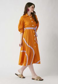 Oranssi nappikiinnitteinen mekko, jossa on puffi hihavat, sisältää purppuran värisiä abstrakteja kuvioita ja siihen kuuluva vyö. Yhdistetty beigeihin liukuvaan sandaaleihin.