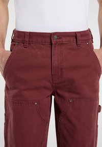 Pantaloni cargo in cotone bordeaux con vita, tasche frontali e accessori decorativi; presentano una superficie testurizzata e cuciture rinforzate.