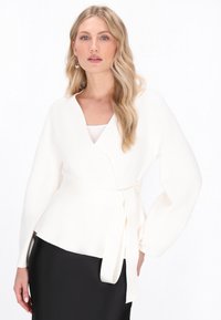 Biała bluzka w stylu wrap, z długimi rękawami i paskiem do wiązania, wykonana z gładkiego materiału. Ma dekolt w kształcie litery V i rozkloszowany dół.