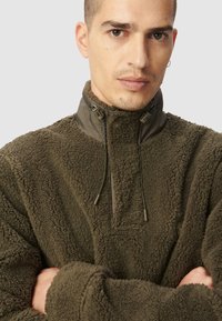Pull-over en polaire vert olive avec un col montant, doté d'une fermeture éclair quart et de cordons de serrage ajustables. Tissu texturé avec une finition douce et moelleuse.