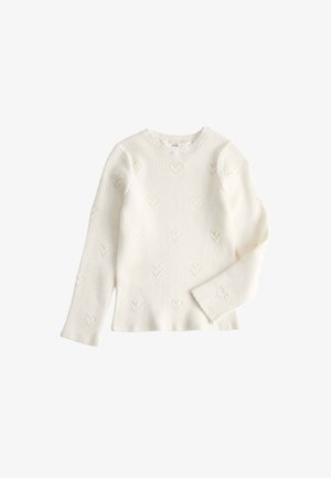 Smilende barn iført en hvid tekstureret sweater og blå jeans med "kindness" broderet på lommen, poserer foran en ensfarvet baggrund.
