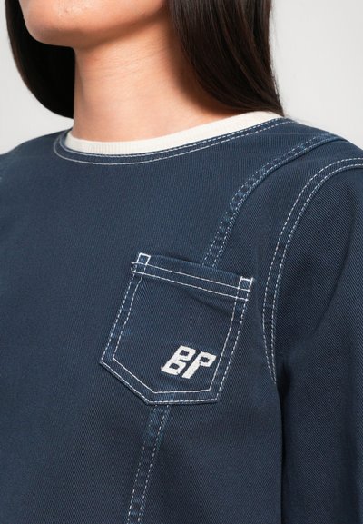 Haut en denim bleu foncé avec un col côtelé blanc, des accents de couture blanche et une poche sur la poitrine gauche avec "BP" brodé en blanc.