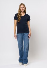 Marinblå t-shirt med rund halsringning och korta ärmar, i kombination med ljusblå utsvängda jeans och vita sneakers. Har en liten logotyp på bröstet.