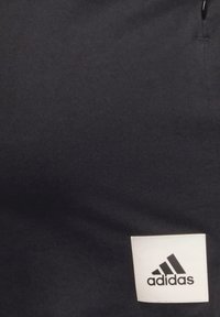 Juodas sportinis švarkas su baltu Adidas logotipo pleistru kairėje pusėje. Medžiaga yra minkšta su lygiu paviršiumi.