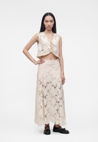 Jeune femme portant un crop top en dentelle crème sans manches et une longue jupe assortie, debout avec une main sur la hanche et des chaussures noires.
