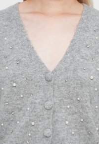 Cardigan gris avec un col en V, présentant des embellissements en perles et strass dispersés, ainsi que trois boutons recouverts de tissu. Texture en maille douce.