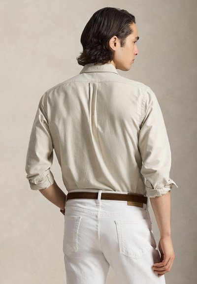 Man met donker haar, opgerolde beige lange-mouwen blouse, bruine riem en witte broek, met de rug naar een neutrale achtergrond gekeerd.