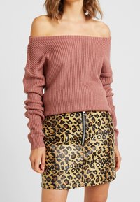 Femme portant un pull en tricot rose poussiéreux à épaules dénudées et une jupe taille haute imprimé léopard avec fermeture éclair à l'avant, sur un fond uni.