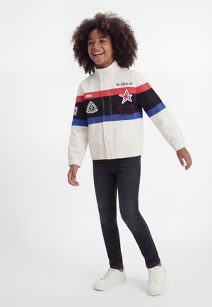 Enfant souriant aux cheveux bouclés portant une veste blanche avec des rayures rouges, noires et bleues, un jean foncé et des baskets blanches, debout sur un fond blanc.