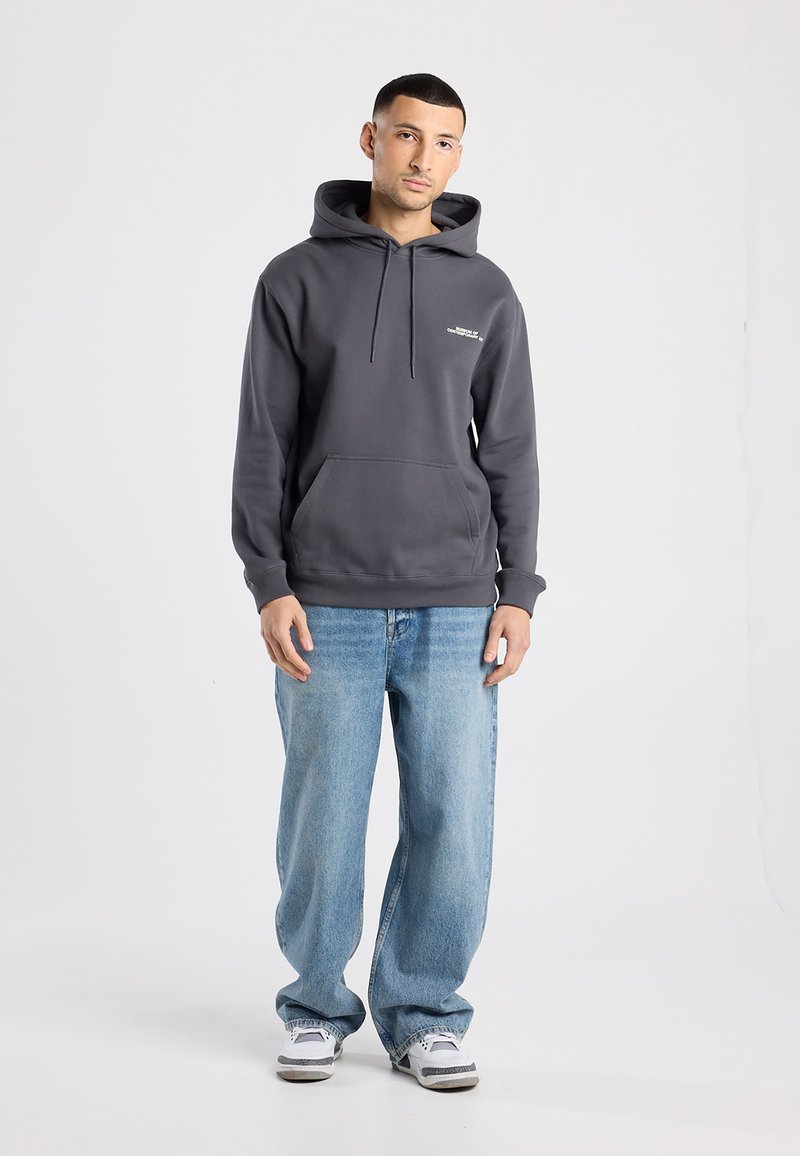 Grijze hoodie van een katoenmix, met kangoeroezak en trekkoord. Gecombineerd met lichtblauwe spijkerbroek en grijze/witte sneakers. Neutrale achtergrond.