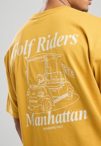 Yellow cotton T-shirt featuring a white graphic of a golf cart and text: "Golf Riders Manhattan Members Only." Simple and casual design.