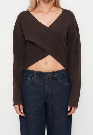 Kvinde iført en kort mørkebrun omslagsstriktrøje med lange ærmer og højtaljede mørkeblå jeans.