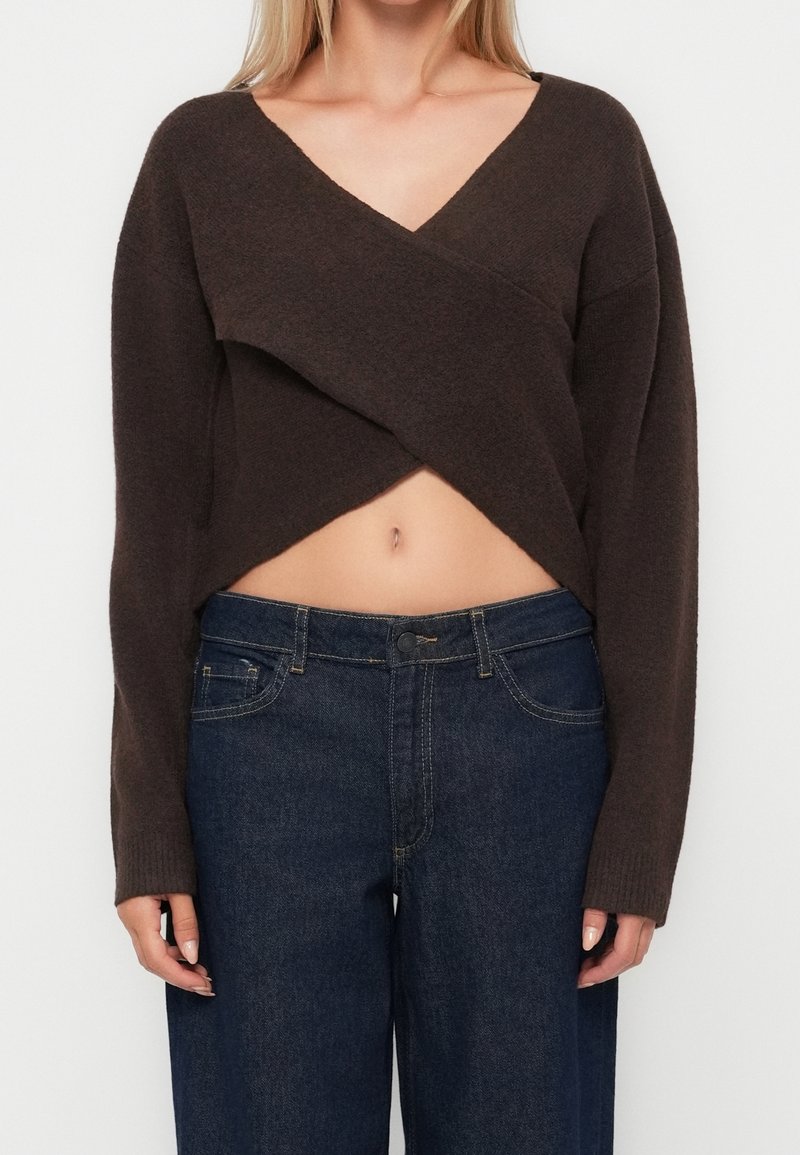 Femme portant un pull court style cache-cœur marron foncé à manches longues et un jean taille haute bleu foncé.