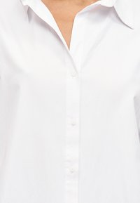 Λευκό πουκάμισο με γιακά και κουμπιά μπροστά, φορεμένο από ένα άτομο με ανοιχτόχρωμο δέρμα ορατό στο λαιμό.