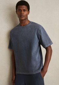 Sivý, prehnaný crewneck tričko vyrobené z mäkkej bavlny s vyblednutým, opotrebovaným povrchom. Krátke rukávy a voľný strih, minimalizované švy.