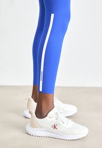 Leggings bleues avec une bande verticale blanche et le texte Calvin Klein, associées à des baskets en mesh blanches Calvin Klein sur un sol clair.