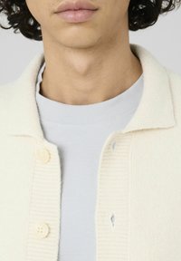 Cardigan en crème avec texture côtelée, col arrondi et quatre boutons. Porté par-dessus un t-shirt gris clair à col rond avec une finition lisse.