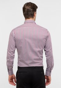Mannen achteraanzicht van een getailleerd shirt met verticale rode en witte strepen, lange mouwen, knopenmanchetten en klassieke kraag; gecombineerd met zwarte broek.