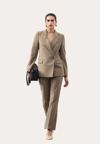 Mujer caminando con un traje de pantalón beige a medida, tacones blancos, pendientes de oro y sosteniendo un bolso de cuero negro contra un fondo liso.