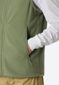 Grön fleeceväst med ståkrage, framzipper och sidoficka. Bärs över en vit långärmad skjorta och tanfärgade byxor.