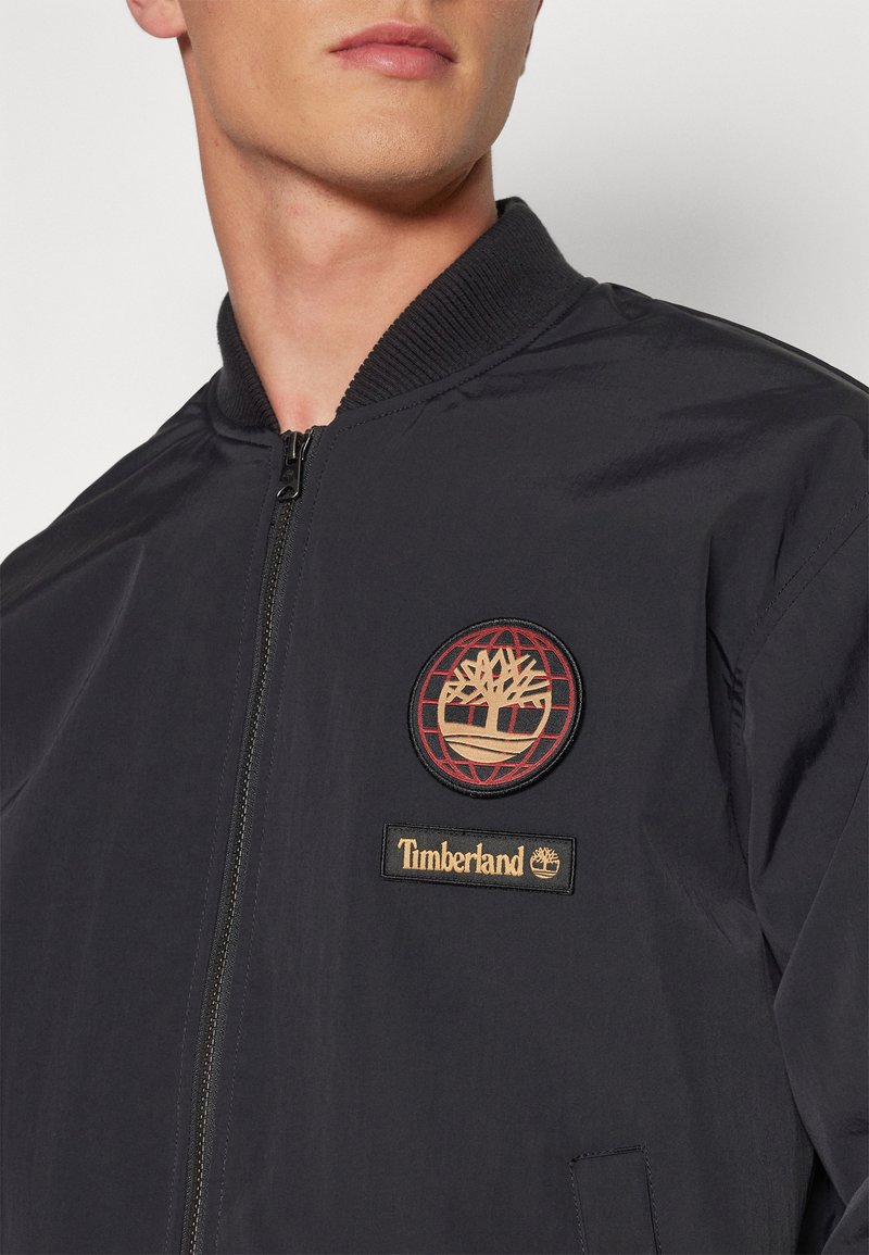 Czarna kurtka bombowa z prążkowanym kołnierzem. Posiada okrągły haftowany logo oraz metkę z napisem "Timberland" w kolorze złotym z przodu.