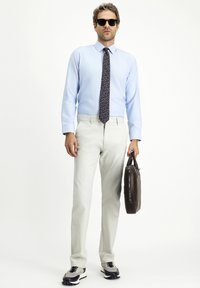 Camicia azzurro chiaro, cravatta a fantasia, pantaloni beige chiaro e occhiali da sole neri. Tiene una valigetta marrone scuro e indossa sneakers grigie e nere.