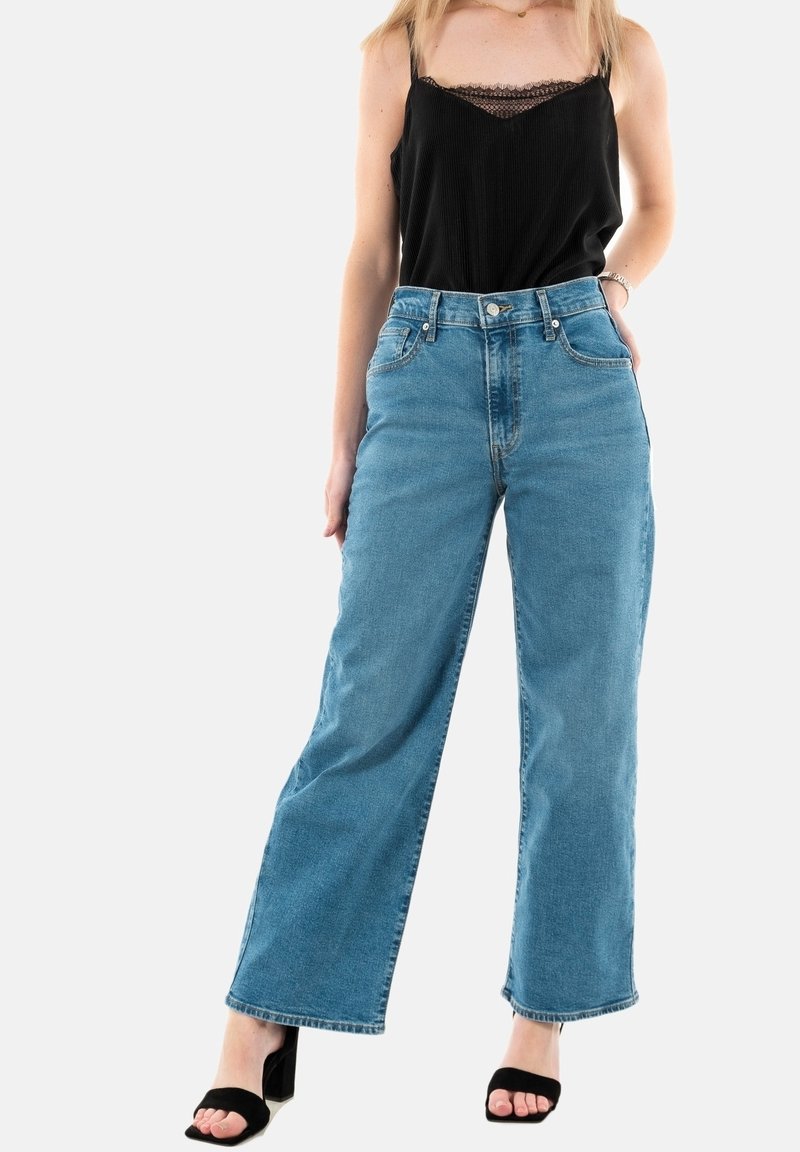 Femme portant un caraco noir avec bordure en dentelle, un jean bleu taille haute à jambes larges et des talons noirs ouverts aux orteils, debout devant un fond uni.
