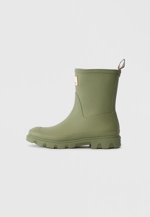 Green rubber ankle boot with textured sole, raised edges, and a pull tab. Features a square label with outlined border. Smooth finish.