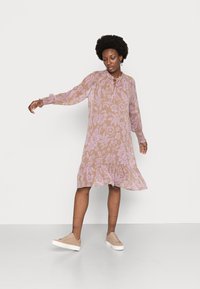 Vestido fluido bege com padrão floral roxo, mangas longas, punhos franzidos e bainha em camadas. Usado com ténis de cor clara.