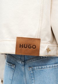 Κοντινό πλάνο προσώπου που φοράει ανοιχτό μπλε τζιν και λευκό μπουφάν με καφέ δερμάτινο ταμπελάκι με την ένδειξη "HUGO" στο κάτω μέρος της πλάτης του μπουφάν.