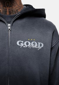 Hoodie nera con zip in tessuto morbido, con testo ricamato "GOOD For Nothing" e tre stelle gialle sul petto.