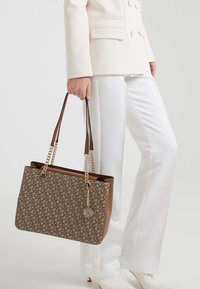 Hnědá vzorovaná taška přes rameno s řetízkovými popruhy, která má zlatý přívěsek. Taška má strukturovaný obdélníkový tvar a hladkou texturu.