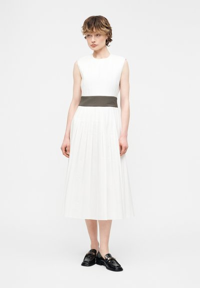 Νεαρή γυναίκα που φοράει αμάνικο λευκό πλισέ φόρεμα με σκούρο ζωνάρι στη μέση και μαύρα μοκασίνια, στέκεται μπροστά σε απλό φόντο.