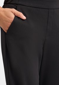 Zwarte broek gemaakt van een gladde, rekbare stof, met een zijzak en strakke, op maat gemaakte naden. De tailleband ligt vlak tegen het lichaam.