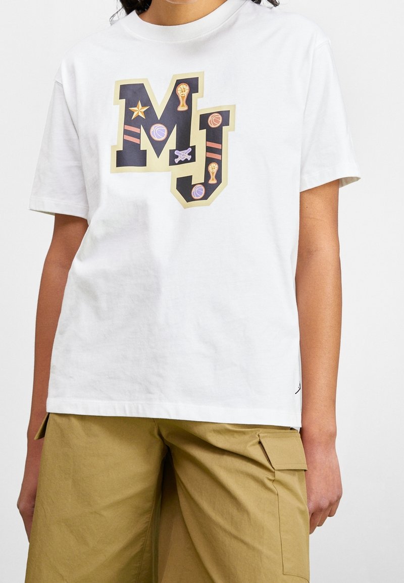 Personne portant un t-shirt blanc avec de grandes lettres "MJ" décorées de ballons de basket, trophées et étoiles, associé à un short cargo beige.