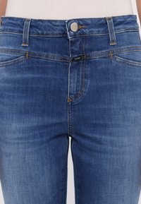 Gros plan sur un jean bleu taille mi-haute avec fermeture par boutons, passants de ceinture et coutures orange contrastantes.