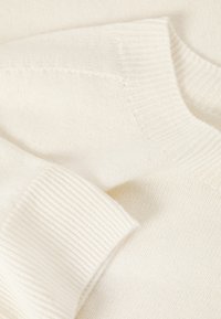 Pull en maille blanche présentant des poignets et un col côtelés avec une texture douce. Le tissu semble léger et lisse.