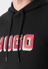 Felpa nera con tasca a marsupio, caratterizzata da una stampa audace del logo "HUGO" in rosso e verde sul davanti. Tessuto testurizzato con lacci regolabili.