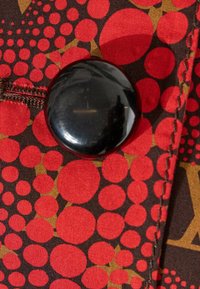 Grand bouton noir brillant cousu sur un tissu avec un fond à motifs circulaires rouges et bruns et des coutures noires visibles.