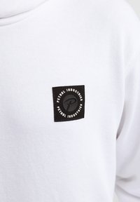 Sweat-shirt blanc avec un patch carré noir comportant le texte et le logo "Petrol Industries". Tissu doux, coupe décontractée, design minimaliste.