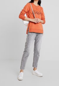 Sudadera naranja con texto negro, corte relajado, con aberturas laterales, combinada con jeans gris claro y zapatillas blancas. Bolso de hombro sencillo en blanco.