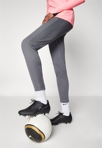 Pantaloni atletici gri cu detalii texturate pe laterale, asortați cu ghete negre, stând pe o minge de fotbal albă și aurie, pe un fundal neutru.