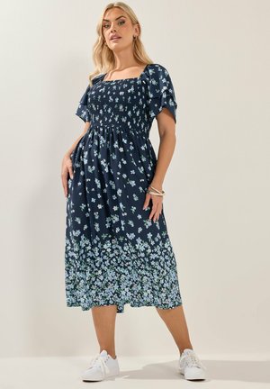 Kvinde iført en navyblå blomsterprint midi-kjole med puffede ærmer og hvide sneakers, stående mod en ensfarvet lys baggrund.