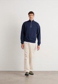 Sudadera de cremallera azul marino con cuello alto, pantalones beiges y zapatos verde oscuro. El modelo está de pie sobre un fondo neutro.