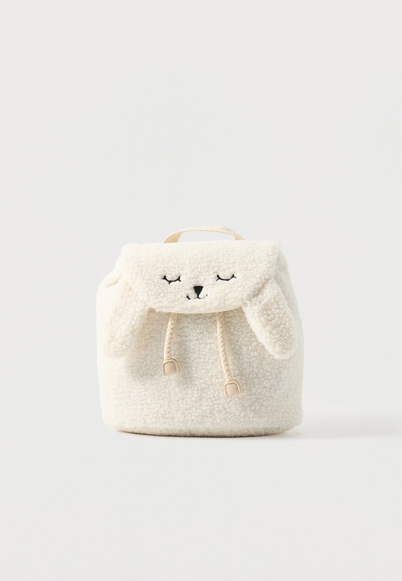 Petit sac à dos blanc duveteux avec fermeture à cordon, rabat avec yeux fermés et nez brodés, et oreilles douces et tombantes sur le rabat.