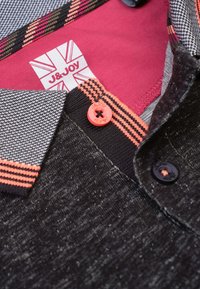 Polo noir texturé avec des accents orange et rose, col rayé, bouton orange et une étiquette intérieure rouge avec « J&Joy ». 